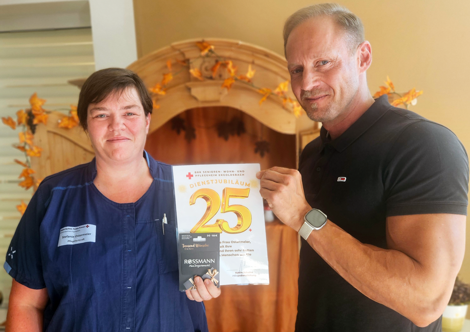 Stefanie Ostermeier Auf dem Bild sind Stefanie (links) und Heimleiter Florian Nahler (rechts) auf dem Bild zu sehen. Stefanie trägt Dienstkleidung mit einem dunkelblauen Oberteil und hält stolz ihre 25-Jahre-Ehrenurkunde in die Kamera. Daneben steht der Heimleiter, Florian Nahler, in einem schwarzen T-Shirt, und hält ebenfalls die Urkunde an einer Ecke, er lächelt voller Anerkennung in die Kamera.