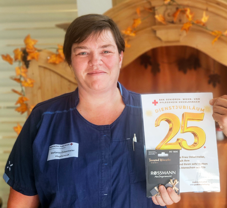 Auf dem Bild sind Stefanie (links) und Heimleiter Florian Nahler (rechts) auf dem Bild zu sehen. Stefanie trägt Dienstkleidung mit einem dunkelblauen Oberteil und hält stolz ihre 25-Jahre-Ehrenurkunde in die Kamera. Daneben steht der Heimleiter, Florian Nahler, in einem schwarzen T-Shirt, und hält ebenfalls die Urkunde an einer Ecke, er lächelt voller Anerkennung in die Kamera.