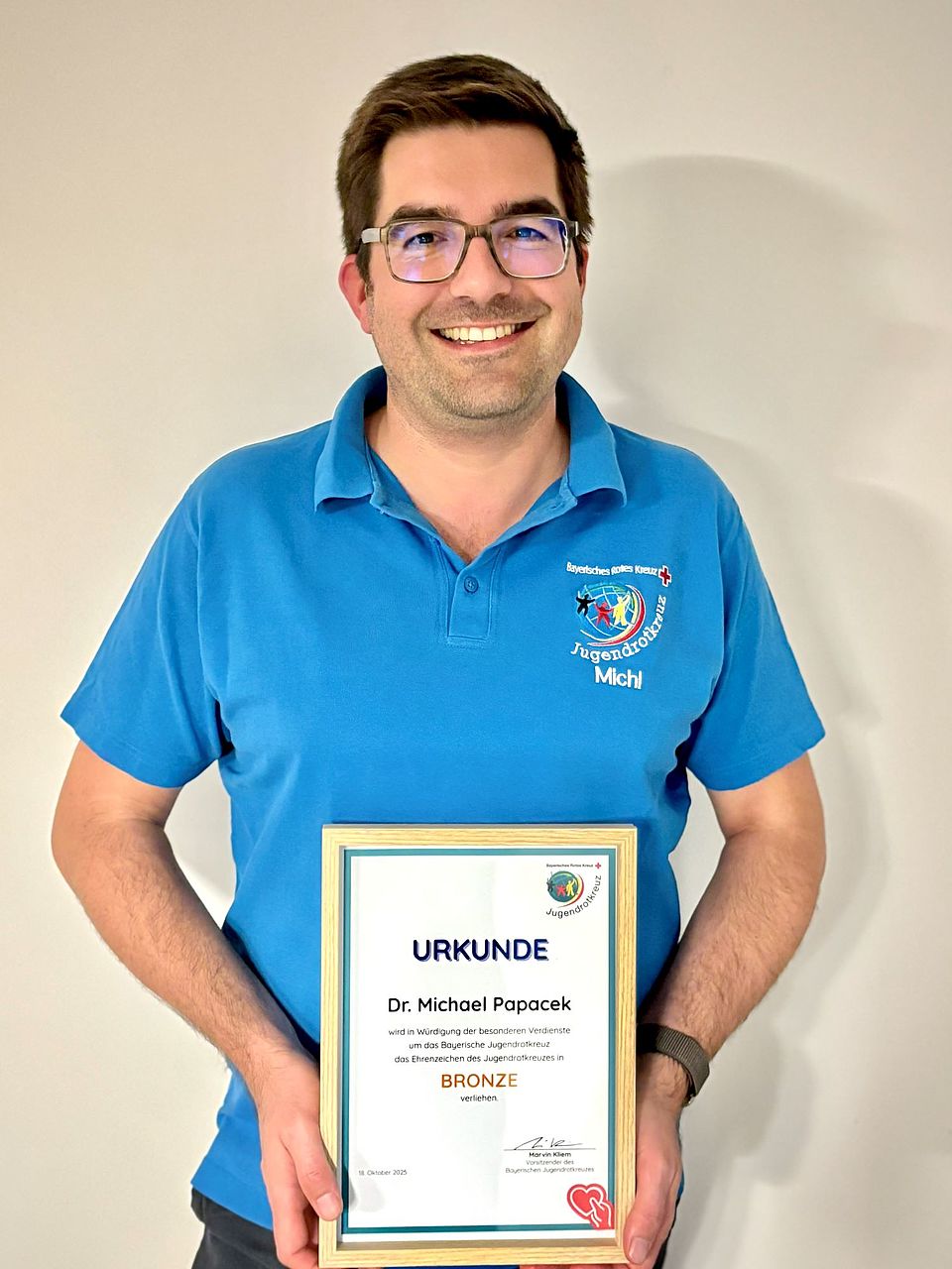 Dr. Michael Papacek Auf dem Bild ist ein Porträt von Dr. Michael Papacek zu sehen. Eine Aufnahme ab Oberkörper. Er hat ein blaues Shirt mit dem Logo des JRK an. Er hält die Ehrenurkunde Bronze in der Hand und lächelt stolz in die Kamera.
