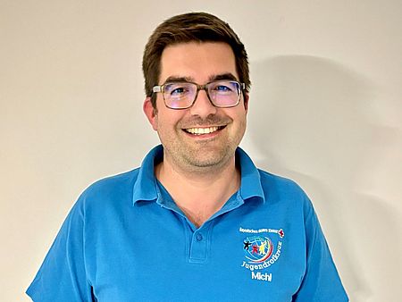 Auf dem Bild ist ein Porträt von Dr. Michael Papacek zu sehen. Eine Aufnahme ab Oberkörper. Er hat ein blaues Shirt mit dem Logo des JRK an. Er hält die Ehrenurkunde Bronze in der Hand und lächelt stolz in die Kamera.