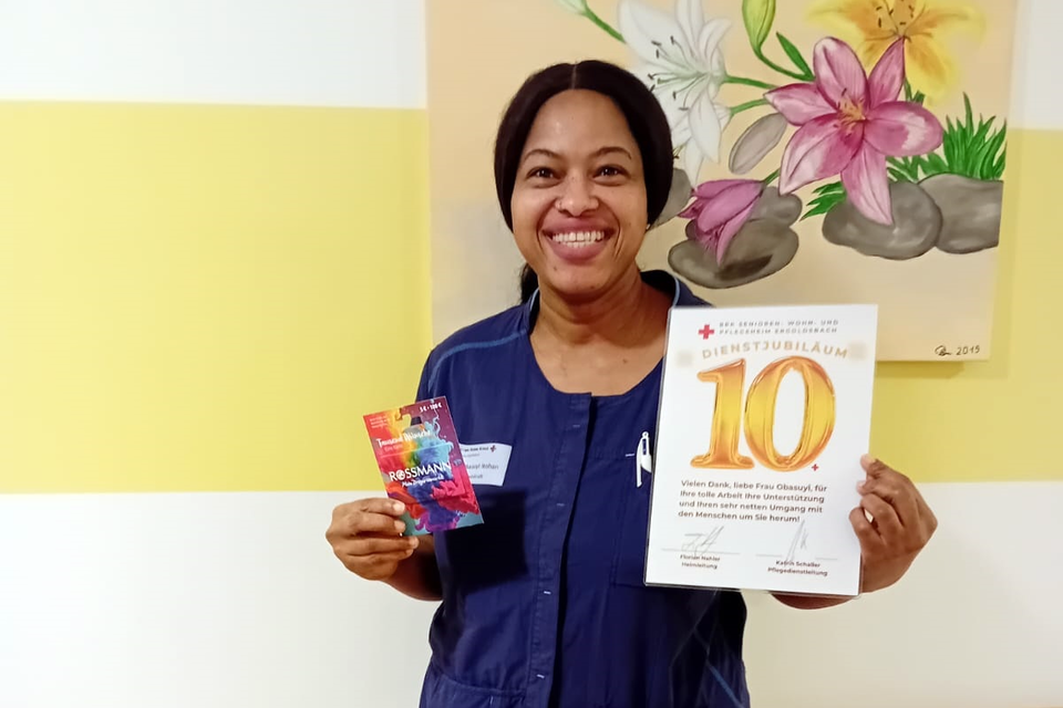 Sarah Obasyui Auf dem Bild ist Sarah in Dienstkleidung zu sehen, weiße Hohe und blaues Oberteil. Sie hält stolz ihre Urkunde in der einen Hand und einen Rossmann-Gutschein in der anderen Hand. Sie lächelt in die Kamera. Der Hintergrund ist eine gelb-weiße Wand mit einem Blümchenbild.