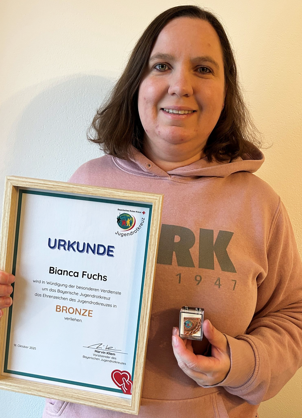 Zu sehen ist Bianca Fuchs als Porträtaufnahme vor einem neutralen hintergrund. Sie hält ihre Ehrenurkunde in der Hand, trägt einen altrosanen JRK-Pullover und lächelt in die Kamera.