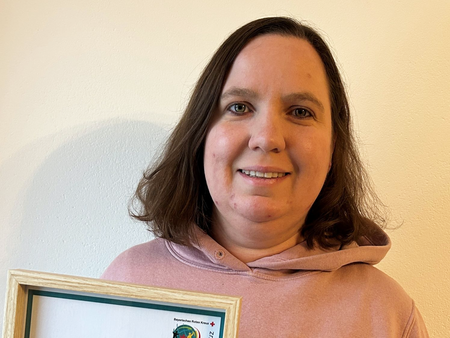 Zu sehen ist Bianca Fuchs als Porträtaufnahme vor einem neutralen hintergrund. Sie hält ihre Ehrenurkunde in der Hand, trägt einen altrosanen JRK-Pullover und lächelt in die Kamera.